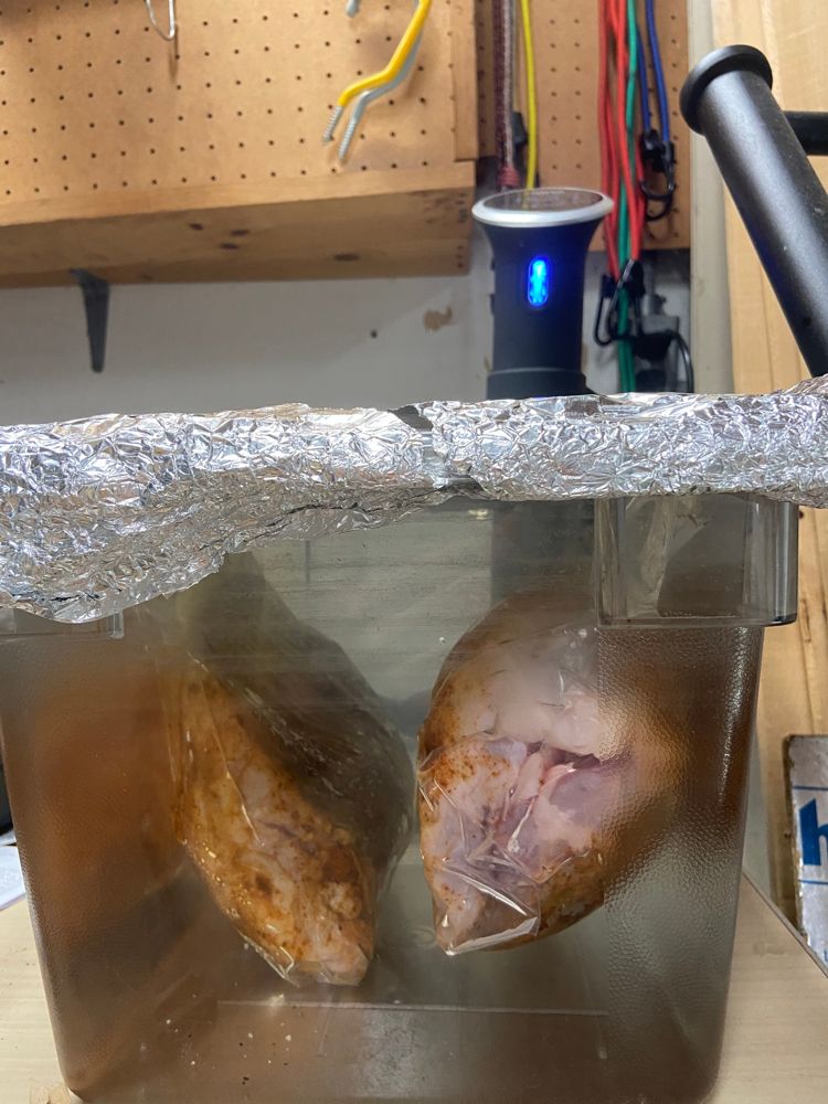 Two bags of seasoned chicken pieces submerged in a bath under a foil lid