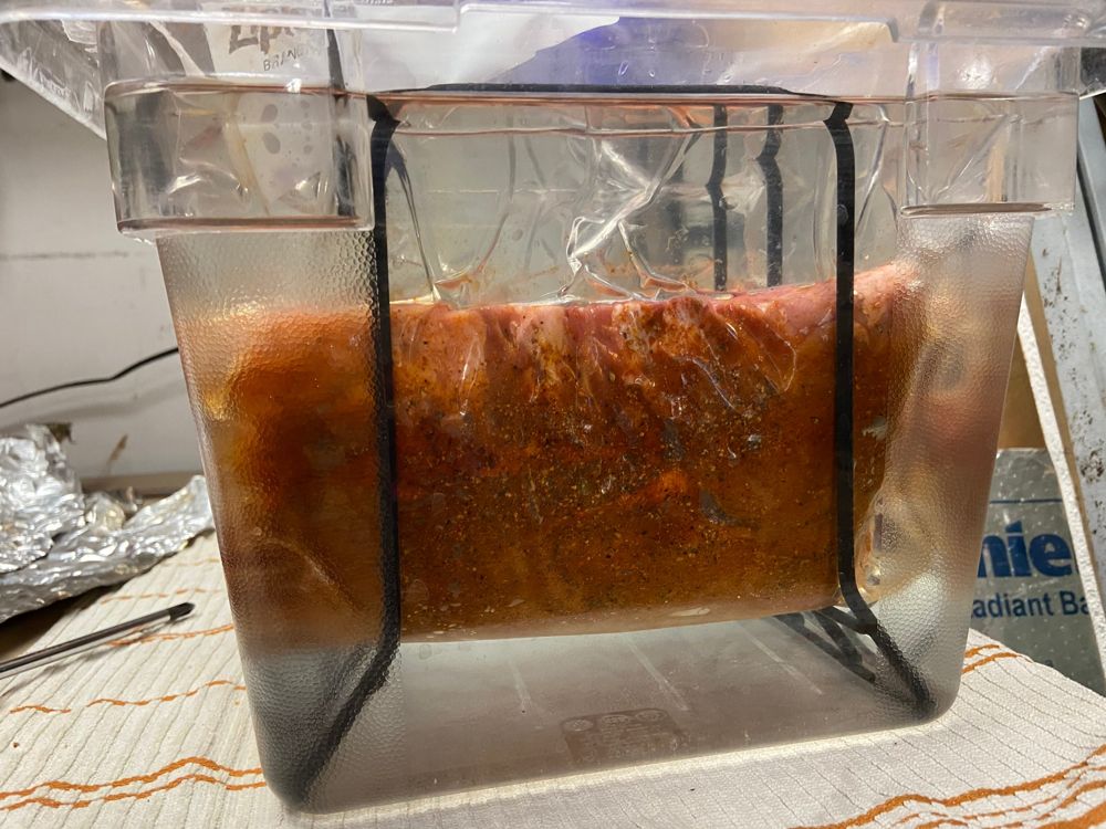 A seasoned rack of meat in a bag held upright in a bath by a rack