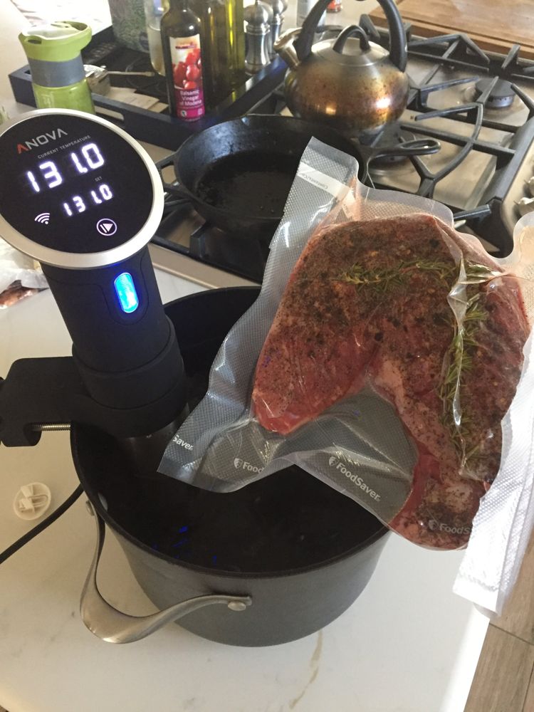 A vacuum-sealed seasoned steak next to a sous vide circulator whose display reads 131.0°F